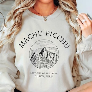 Peut inclure: Sweat-shirt couleur sable avec un dessin noir de Machu Picchu, la Cité perdue des Incas, Cusco, Pérou. Le texte "Machu Picchu" est arqué au-dessus de l'image, et les mots "Little Swan Crafts" sont sur le côté gauche.
