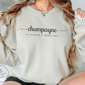 May include: Sand-colored sweatshirt with the text "champagne is always a good idea" in black script. The sweatshirt has long sleeves and a crew neck. The brand logo "Little Swan Crafts" is visible on the lower left.
