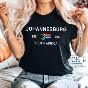 May include: Black t-shirt with white text that reads "JOHANNESBURG EST. 1886 SOUTH AFRICA" and a small image of the South African flag.