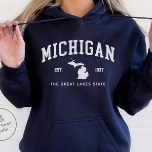 May include: Navy blue hoodie with the word "MICHIGAN" in white block letters. Below the word "MICHIGAN" is the text "EST. 1837" and a white outline of the state of Michigan. The text "THE GREAT LAKES STATE" is below the state outline.