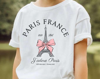 May include: White t-shirt featuring a graphic of the Eiffel Tower with a pink bow, the words "Paris France" and "J'adore Paris". The shirt is worn by a person and paired with denim shorts. The shirt design includes the text "RÉPUBLIQUE FRANÇAISE".