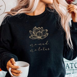Puede incluir: Una sudadera negra de manga larga con cuello redondo que presenta una ilustración de líneas doradas de una flor de loto centrada en el pecho, con el texto "no mud no lotus" impreso en una fuente cursiva dorada bajo el gráfico.