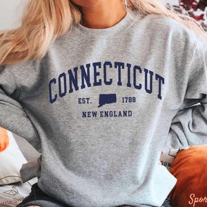 Peut inclure: Un sweat-shirt gris avec le mot "CONNECTICUT" en lettres majuscules bleu marine. En dessous, on peut lire "EST. 1788" et "NEW ENGLAND". Le sweat-shirt est de style col rond et est fait d'une matière douce.