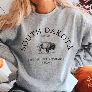 May include: Sport grey sweatshirt with a black graphic of a bison and the words "South Dakota" and "The Mount Rushmore State". The sweatshirt also has the text "Est. 1889". The sweatshirt is a comfortable, casual style.