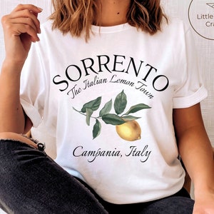May include: White t-shirt with the text "Sorrento The Italian Lemon Town Campania, Italy" in black script. A watercolor illustration of a lemon branch with green leaves and a yellow lemon is below the text. The shirt has rolled-up sleeves.