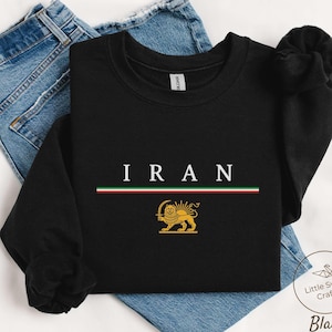 Op de afbeelding: Zwart sweatshirt met lange mouwen met het woord "IRAN" in witte hoofdletters. Daaronder een horizontale streep in rood, wit en groen. Een gouden leeuwenembleem staat onder de streep. Het sweatshirt wordt getoond op een blauwe jeans.