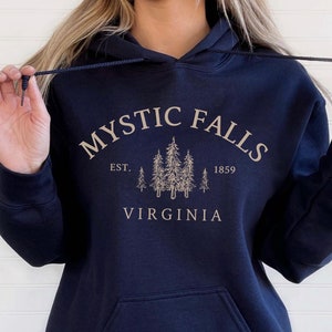 May include: A navy blue hooded sweatshirt featuring a front kangaroo pocket and long sleeves. The chest displays a beige graphic with the text "MYSTIC FALLS", "EST. 1859", and "VIRGINIA" surrounding a detailed illustration of five pine trees.