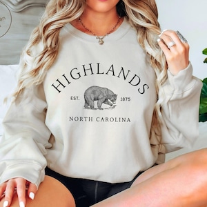 以下が含まれることがあります： 砂色のスウェットシャツ。上部に「HIGHLANDS」、下部に「NORTH CAROLINA」と書かれています。中央にはクマのイラストがあり、その上に「EST. 1875」と書かれています。クルーネックのスタイルのスウェットシャツです。