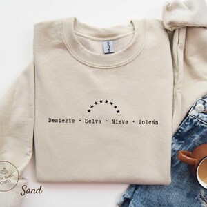 May include: A sand-coloured sweatshirt with a crew neck. The design features a semi-circle of stars above the words "Desierto, Selva, Nieve, Volcán" in black. A small logo and the word "Sand" are visible on the sleeve.