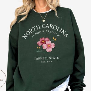 May include: Forest green sweatshirt with the text "NORTH CAROLINA" and coordinates, a floral design, and "TARHEEL STATE EST. 1789". The sweatshirt is a dark green color.