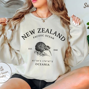 Könnte beinhalten: Ein sandfarbenes Sweatshirt mit einem schwarzen Grafikdruck eines Kivis und dem Text "NEW ZEALAND PACIFIC OCEAN EST. 1840 41°18′S 174°47′E OCEANIA".