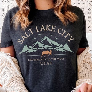 May include: Dark grey heather t-shirt with a graphic design featuring a mountain range, a moose, and the text "Salt Lake City, Crossroads of the West, Utah".