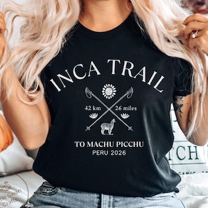 Könnte beinhalten: Ein schwarzes T-Shirt mit weißem Text, der INCA TRAIL TO MACHU PICCHU PERU 2026 lautet. Das Design zeigt zwei gekreuzte Wanderstöcke, ein Sonnensymbol, zwei kleine Blumen und ein Lama, sowie die Angaben 42 km und 26 miles.