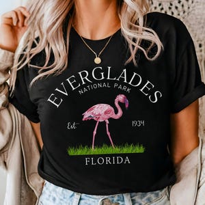 May include: Black t-shirt featuring a white graphic of a pink flamingo in front of green grass. The graphic reads "Everglades National Park Est. 1934 Florida".