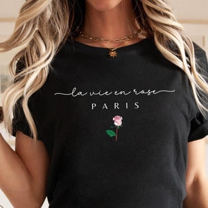 May include: Black t-shirt with the phrase "la vie en rose PARIS" in white script and a pink rose illustration. The shirt is a classic crew neck style.
