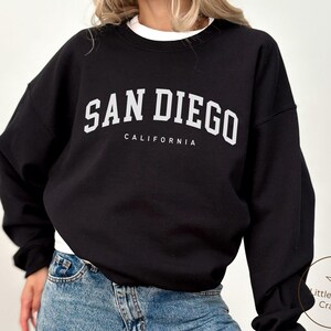 May include: Black sweatshirt with the text "SAN DIEGO CALIFORNIA" in white block letters. The sweatshirt has long sleeves and a crew neck. The model is wearing blue jeans.