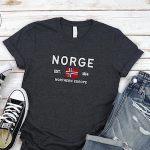 以下が含まれることがあります： ダークグレーヘザーのTシャツに、白い文字で「NORGE EST. 1814 NORTHERN EUROPE」と、ノルウェーの国旗の小さな画像がプリントされています。