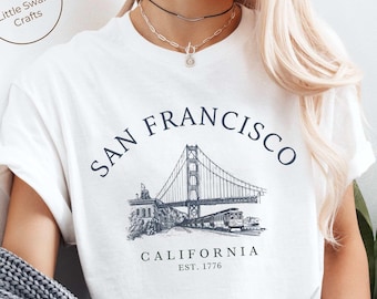 Puede incluir: Camiseta blanca con un gráfico del Puente Golden Gate en San Francisco, California. El texto "SAN FRANCISCO" está escrito en un arco sobre el puente, y el texto "CALIFORNIA EST. 1776" está escrito debajo del puente.