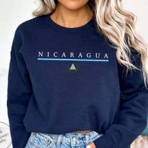 Może przedstawiać: Granatowa bluza z napisem "NICARAGUA" białymi wielkimi literami, nad niebiesko-białym paskiem i małym emblematem. Bluza ma okrągły dekolt.