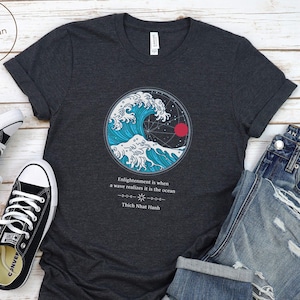 Könnte beinhalten: Dunkelgraues T-Shirt mit einem blauen und weißen Wellen-Motiv und einer roten Sonne. Der Text "Enlightenment is when a wave realizes it is the ocean" und "Thich Nhat Hanh" ist unter dem Motiv gedruckt.