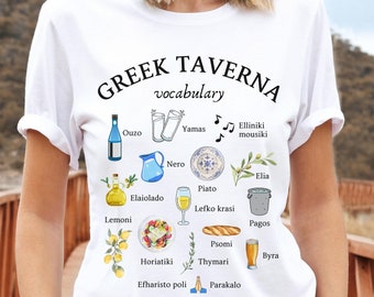 May include: White t-shirt with a graphic design of Greek food and drink vocabulary. The design includes illustrations of olives, lemons, bread, wine, beer, and more. The text includes words like "Ouzo", "Yamas", "Elaiolado", "Horiatiki", "Lefko krasi", "Psomi", "Byra", "Efharisto poli", and "Parakalo".