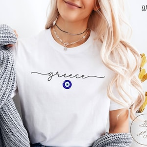 May include: White t-shirt with the word "greece" written in cursive script and a blue evil eye charm below it.
