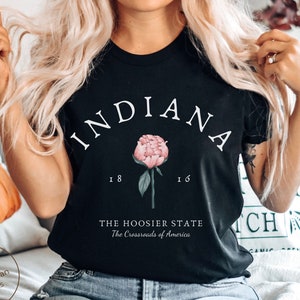 Può includere: Maglietta nera con testo bianco che recita "INDIANA" in un arco curvo con un fiore rosa stilizzato e il testo "THE HOOSIER STATE The Crossroads of America".
