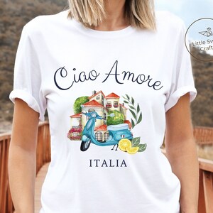 May include: White t-shirt with a blue and white scooter, a small building with red roofs, and green plants. The text on the shirt reads 'Ciao Amore' and 'ITALIA'.