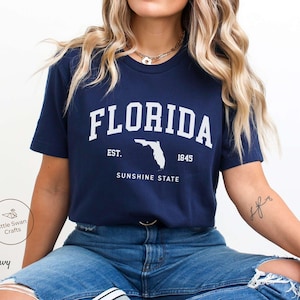 May include: Navy blue t-shirt with the word "FLORIDA" in white block letters, a white outline of the state, and the text "EST. 1845 SUNSHINE STATE". The shirt is a short-sleeved crew neck.