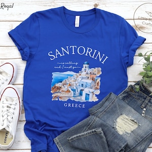 May include: Blue t-shirt with a white graphic of Santorini, Greece. The graphic includes a watercolor illustration of the town and the text "Santorini is calling and I must go".