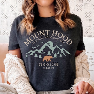 May include: Dark grey heather t-shirt featuring a graphic of Mount Hood, Oregon. The design includes mountains, trees, a bear, and the text "MOUNT HOOD CASCADE VOLCANIC ARC OREGON 11.249 FT". The shirt is a comfortable, casual wear.