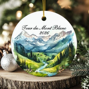 May include: A round white ceramic ornament featuring a watercolor illustration of the Tour du Mont Blanc mountain range, a green valley, a winding blue river, and pine trees, with the text Tour du Mont Blanc 2026 printed in black.