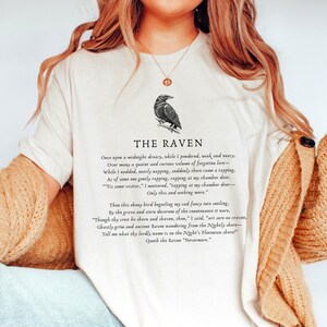 Puede incluir: Una camiseta blanca con una ilustraci&oacute;n de cuervo negro y el texto "THE RAVEN" impreso en la parte delantera. El texto debajo de la ilustraci&oacute;n del cuervo es un extracto del poema "El Cuervo" de Edgar Allan Poe.