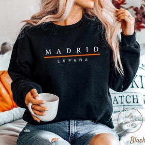 May include: Black crew neck sweatshirt with the word "MADRID" in white capital letters, with a horizontal orange and red stripe underneath. The word "ESPAÑA" is printed below in white. A white mug is held in the hands.