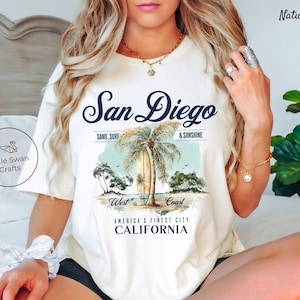 Peut inclure: T-shirt blanc avec un motif graphique bleu et blanc représentant des palmiers, une planche de surf et le texte "San Diego, Sand, Surf & Sunshine, West Coast, America's Finest City, California".