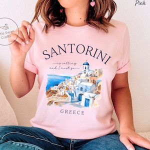 May include: Pink t-shirt with a watercolor illustration of Santorini, Greece. The text on the shirt reads "Santorini is calling and I must go".