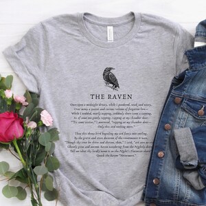 Puede incluir: Una camiseta gris con una ilustraci&oacute;n de cuervo negro y el texto "THE RAVEN" impreso en la parte delantera. El texto debajo del cuervo incluye las primeras l&iacute;neas del poema "El Cuervo" de Edgar Allan Poe.