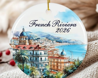 May include: A round white ceramic ornament featuring a watercolour illustration of the French Riviera coastline with buildings, palm trees, and blue water. The text "French Riviera" and "2026" is printed in dark blue script and serif font.