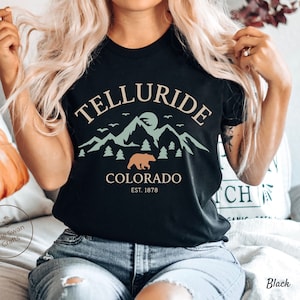 May include: Black t-shirt with a graphic design featuring the word "TELLURIDE" above a mountain scene, a bear, and the word "COLORADO" with "EST. 1878". The design is in shades of green, tan, and white.