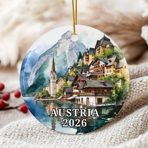May include: A round ceramic ornament featuring a watercolor-style illustration of an Austrian lakeside village with a church, mountains, and houses. The text AUSTRIA 2026 is printed in black at the bottom. It hangs from a thin gold string.