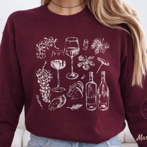 Könnte beinhalten: Burgunderfarbenes Sweatshirt mit einer weißen Illustration von Weinelementen. Das Design umfasst Weingläser, Flaschen, Trauben, einen Korkenzieher und Blätter. Die Wörter "Little Swan Crafts" und "Maroon" sind ebenfalls sichtbar.