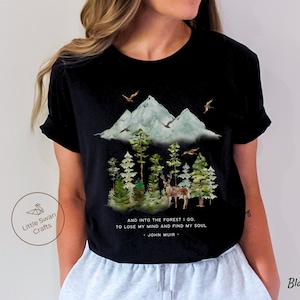 Pode incluir: Camiseta preta com uma ilustra&ccedil;&atilde;o em aquarela de uma cordilheira, &aacute;rvores e um veado. O texto "AND INTO THE FOREST I GO. TO LOSE MY MIND AND FIND MY SOUL - JOHN MUIR -" est&aacute; impresso na camiseta.