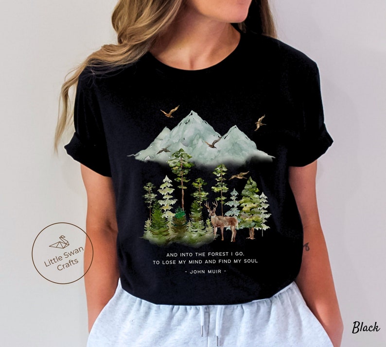 Pode incluir: Camiseta preta com uma ilustra&ccedil;&atilde;o em aquarela de uma cordilheira, &aacute;rvores e um veado. O texto "AND INTO THE FOREST I GO. TO LOSE MY MIND AND FIND MY SOUL - JOHN MUIR -" est&aacute; impresso na camiseta.