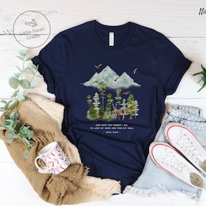 Pode incluir: Camiseta azul marinho com uma ilustra&ccedil;&atilde;o em aquarela de uma cena de floresta com montanhas, &aacute;rvores e um veado. O texto "AND INTO THE FOREST I GO TO LOSE MY MIND AND FIND MY SOUL - JOHN MUIR -" est&aacute; impresso na camiseta.