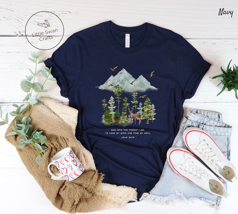 Pode incluir: Camiseta azul marinho com uma ilustra&ccedil;&atilde;o em aquarela de uma cena de floresta com montanhas, &aacute;rvores e um veado. O texto "AND INTO THE FOREST I GO TO LOSE MY MIND AND FIND MY SOUL - JOHN MUIR -" est&aacute; impresso na camiseta.