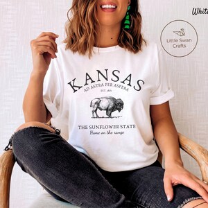 May include: White t-shirt with a black graphic design featuring the text "KANSAS", "AD ASTRA PER ASPERA", "EST. 1861", "THE SUNFLOWER STATE", and "Home on the range". The graphic also includes a black and white illustration of a buffalo.