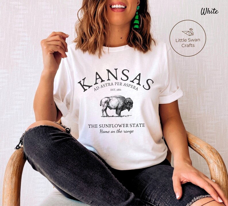 May include: White t-shirt with a black graphic design featuring the text "KANSAS", "AD ASTRA PER ASPERA", "EST. 1861", "THE SUNFLOWER STATE", and "Home on the range". The graphic also includes a black and white illustration of a buffalo.
