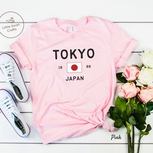 K&ouml;nnte beinhalten: Rosa T-Shirt mit dem Text "TOKYO" und der japanischen Flagge. Der Text "18" und "68" befindet sich auf beiden Seiten der Flagge. Der Text "JAPAN" befindet sich unter der Flagge.