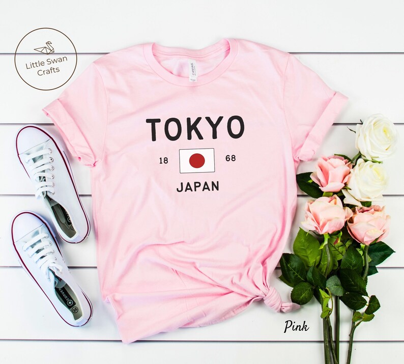 K&ouml;nnte beinhalten: Rosa T-Shirt mit dem Text "TOKYO" und der japanischen Flagge. Der Text "18" und "68" befindet sich auf beiden Seiten der Flagge. Der Text "JAPAN" befindet sich unter der Flagge.