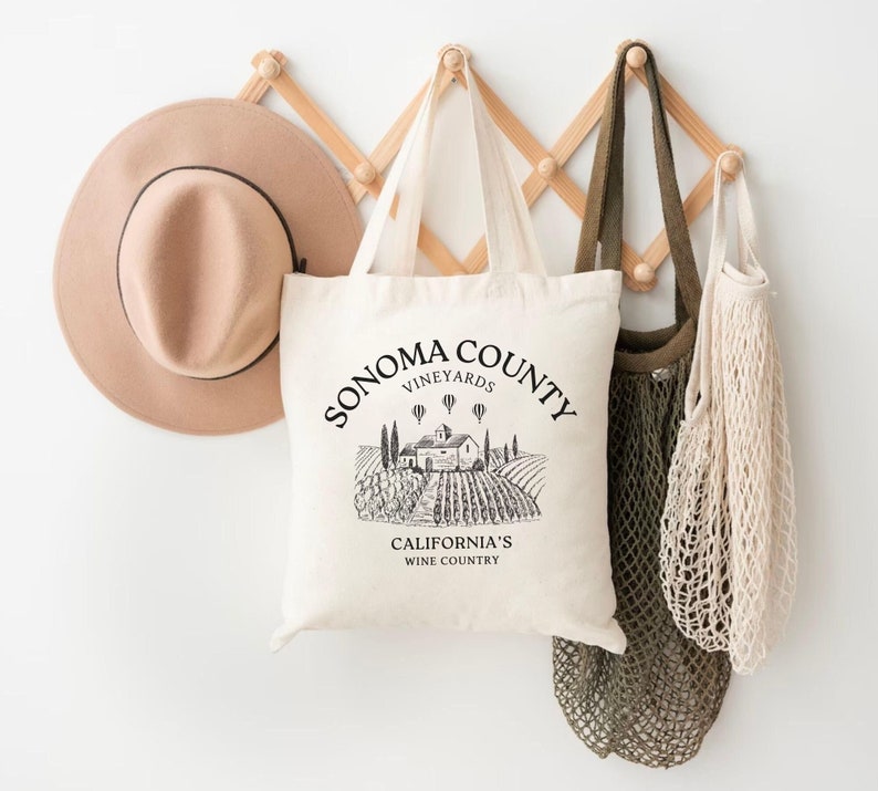 May include: A white tote bag with black text that reads "Sonoma County Vineyards California's Wine Country". The bag has a black and white illustration of a vineyard with a house and hot air balloons. The bag is hanging on a wooden rack with a brown hat and two string bags.